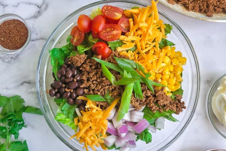 a taco salad in a large bowl with lettuce, corn, beans, onions, tomatoes, cheese, ground beef, tortilla chips. Sour cream, salsa, cilantro, lime wedges and taco seasoning scattered around the salad, skillet of cooked ground beef behind the salad