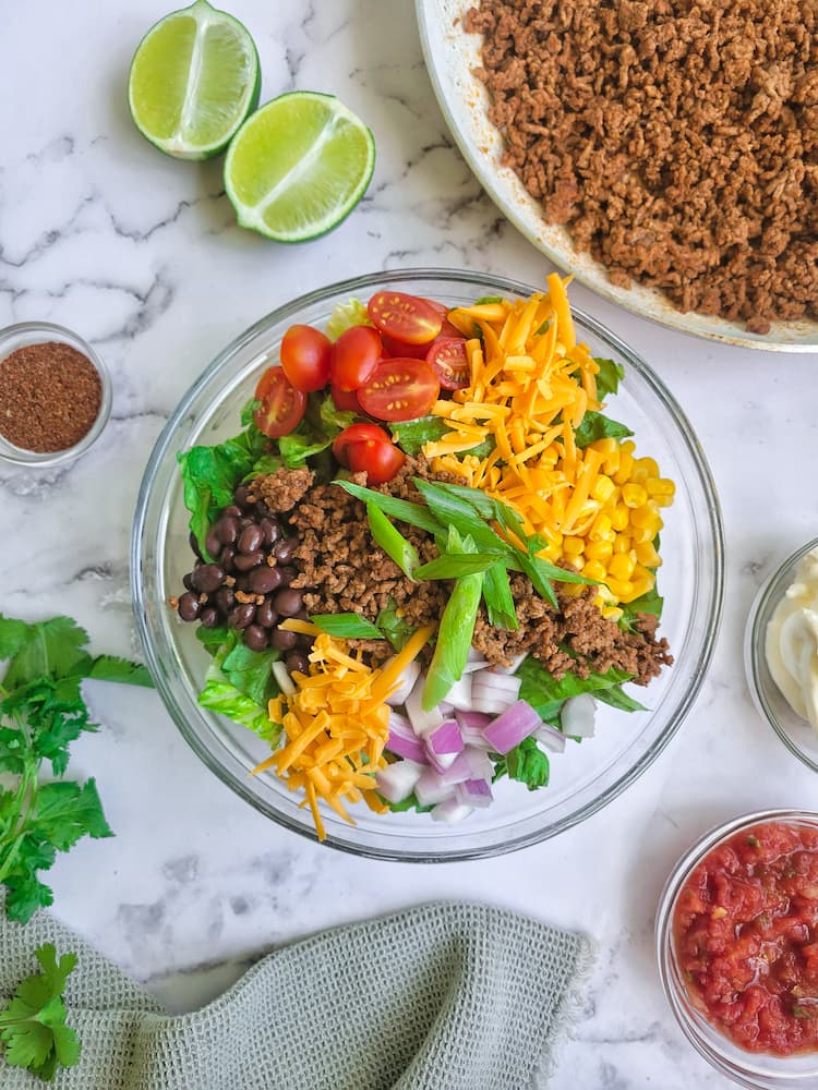 a taco salad in a large bowl with lettuce, corn, beans, onions, tomatoes, cheese, ground beef, tortilla chips. Sour cream, salsa, cilantro, lime wedges and taco seasoning scattered around the salad, skillet of cooked ground beef behind the salad