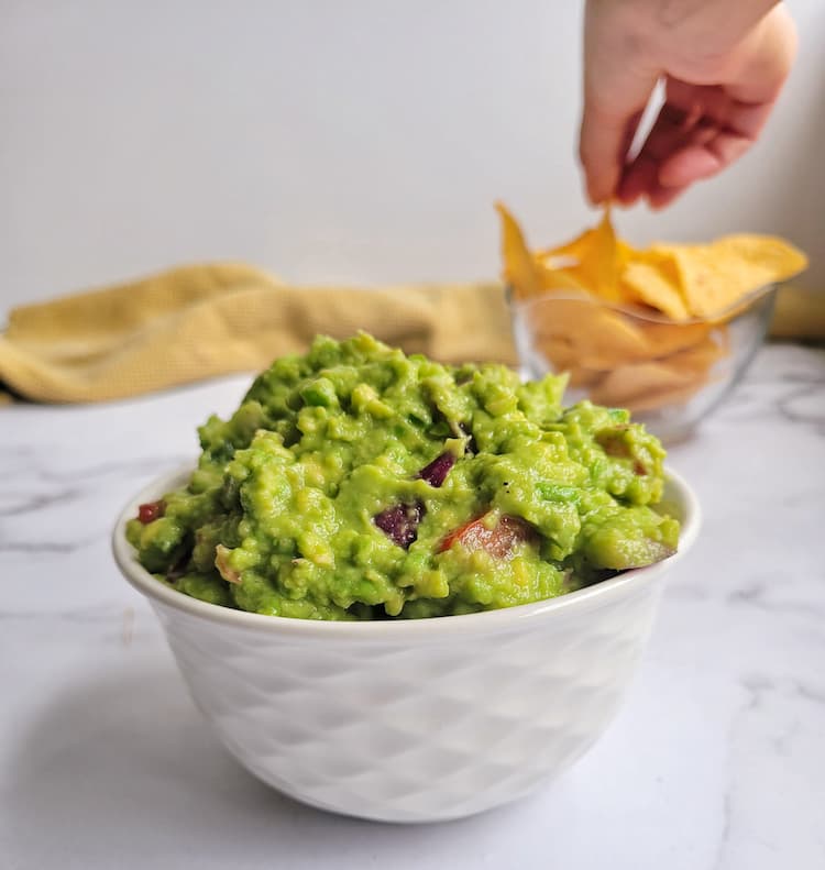 bowl of guacamole, bowl of tortilla chips in the background with hand grabbing one