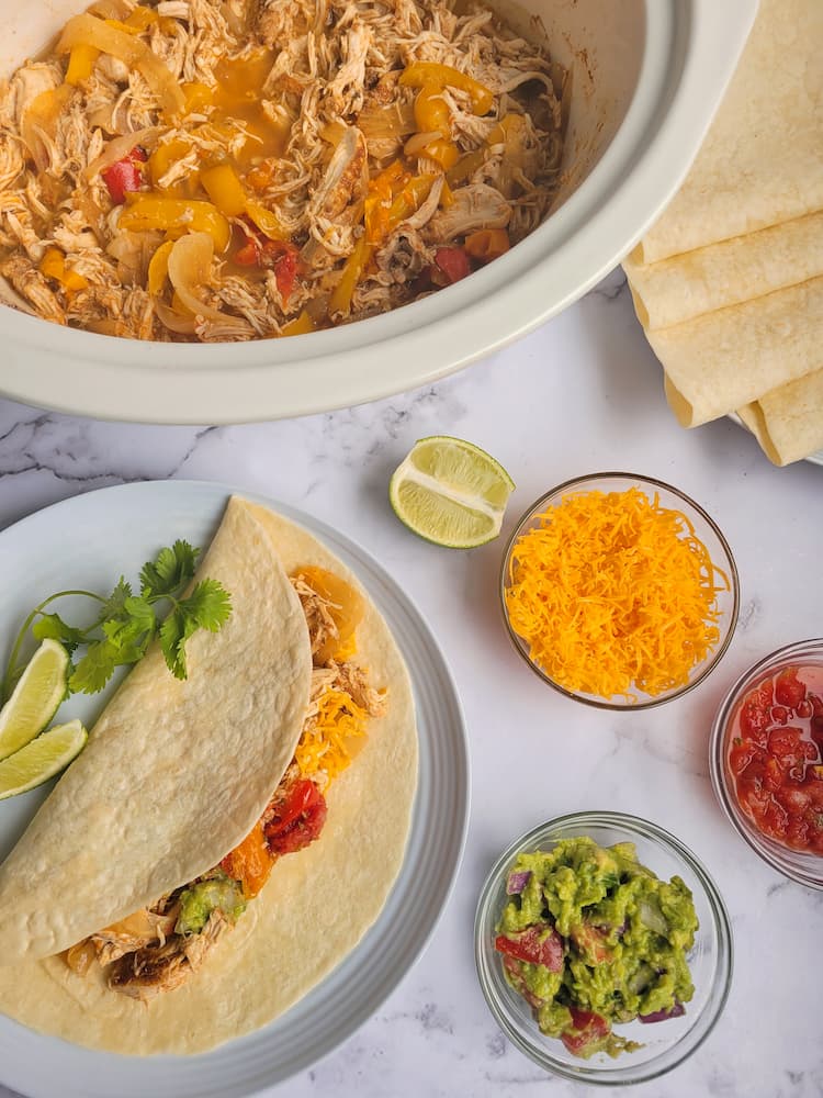 crockpot of shredded cooked chicken and peppers next to a plate with a loaded chicken fajita on it. Bowls of sour cream, salsa and guacamole with a half a lime around, more tortillas on a plate in the background