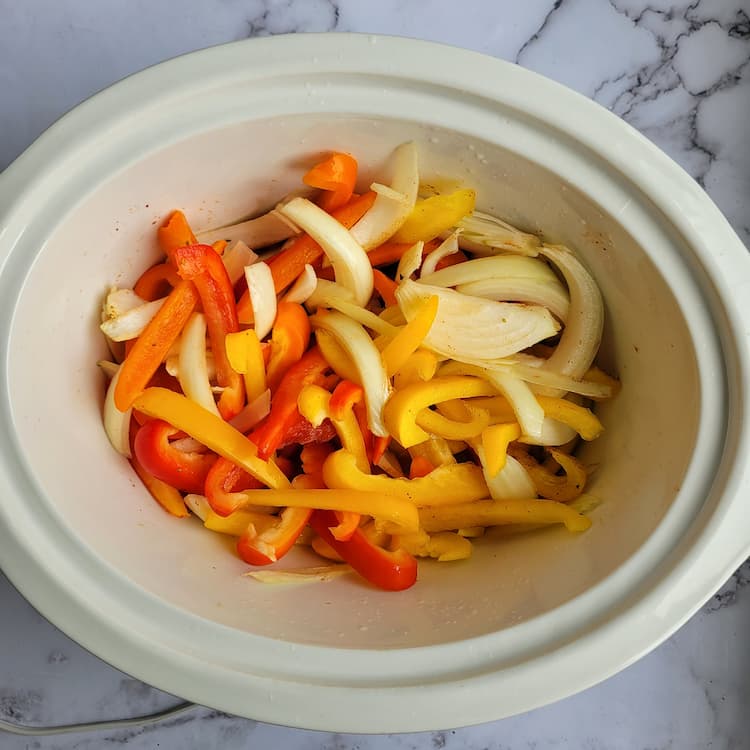 crockpot filled with sliced red, orange and yellow peppers and a white onion