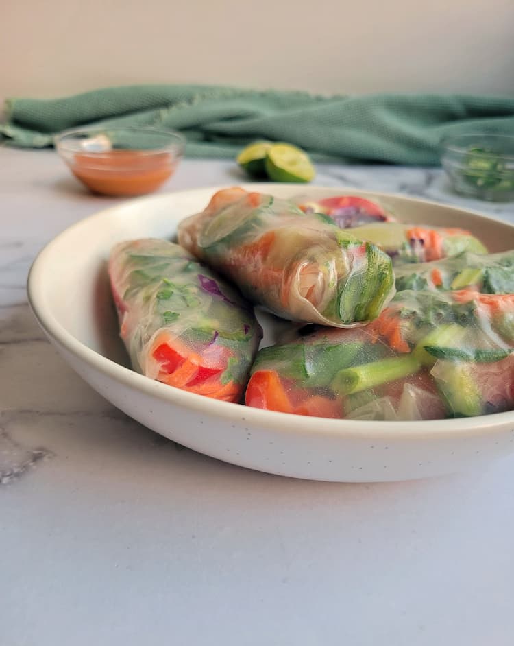side view of a bowl of fresh spring rolls, lime wedge and peanut sauce in the background