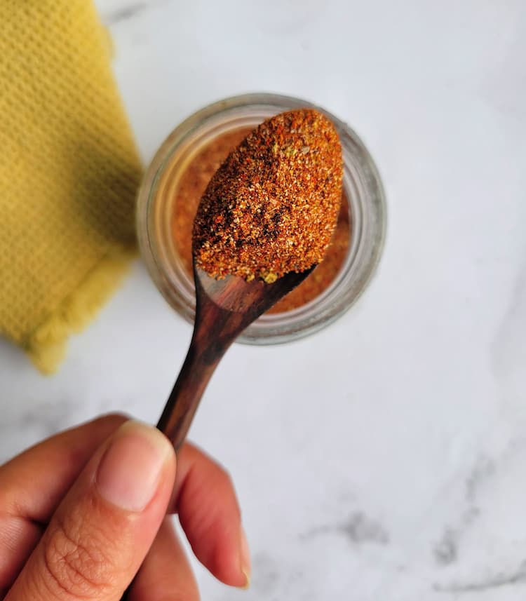 hand holding a wooden spoon with some spices on it over a jar of the rest