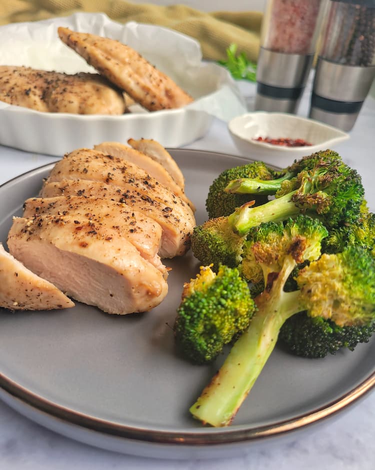 plate with sliced chicken breast and roasted broccoli, extra chicken in the background with salt and pepper mills