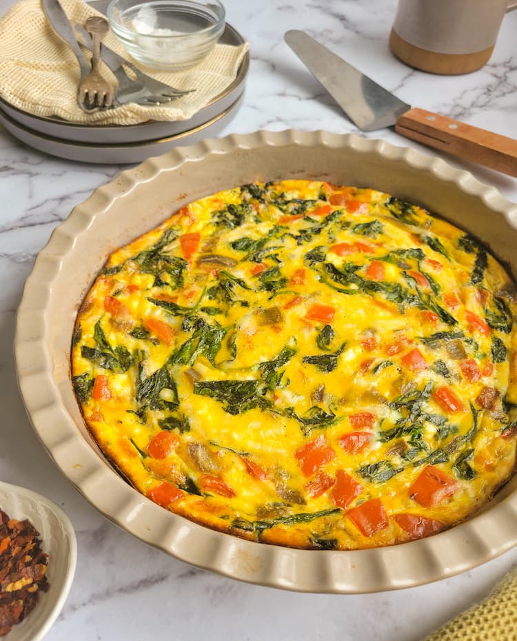 side view of baked quiche with red peppers and spinach in a pie dish, ramekin of chili flakes, cup of coffee, serving spatula and plates with forks around and in the background