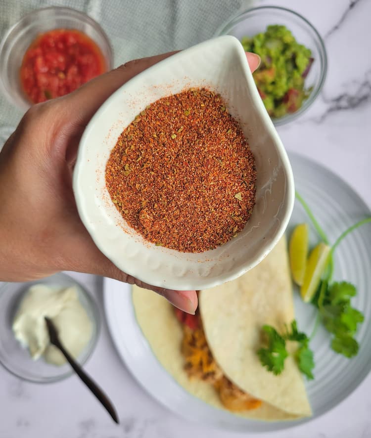 hand holding a small ramekin of spices over a fajitas, and bowls of guacamole, sour cream and salsa