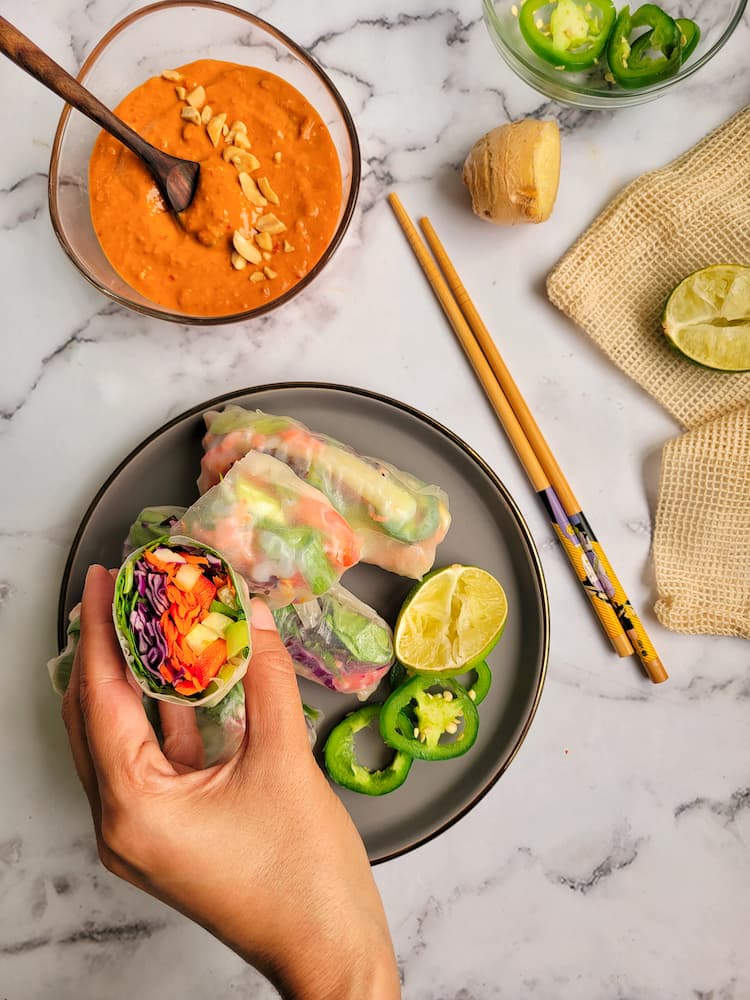 hand holding a cut fresh veggie spring roll, bowl of peanut sauce with chopped peanuts in the background, chopsticks, a knob of ginger, a halved lime and a ramekin of sliced jalapenos