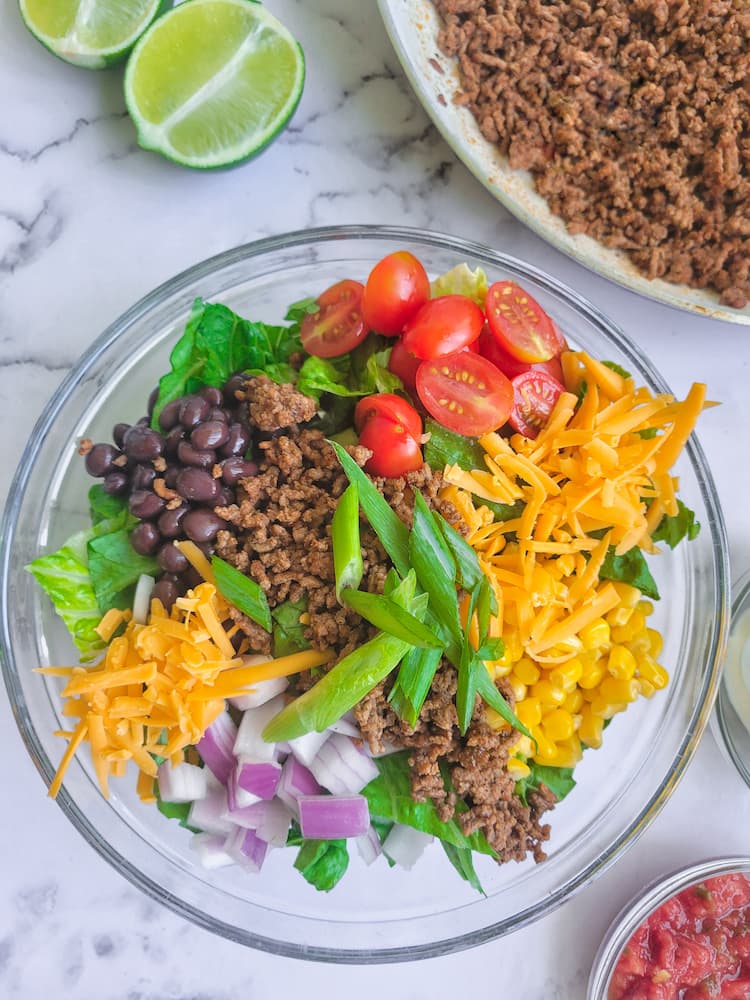 a taco salad in a large bowl with lettuce, corn, beans, onions, tomatoes, cheese, ground beef. Skillet of ground beef in the background with halved lime wedges, small bowl of salsa