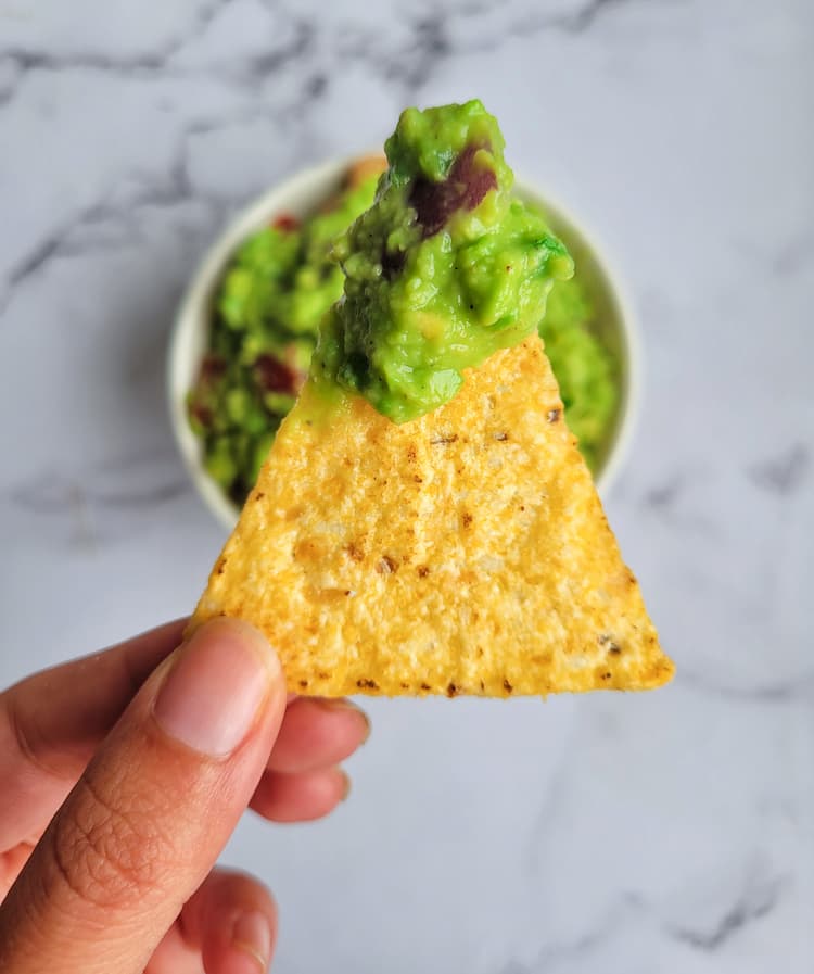 hand holding up a tortilla chip with some guacamole on it, over a bowl of guacamole