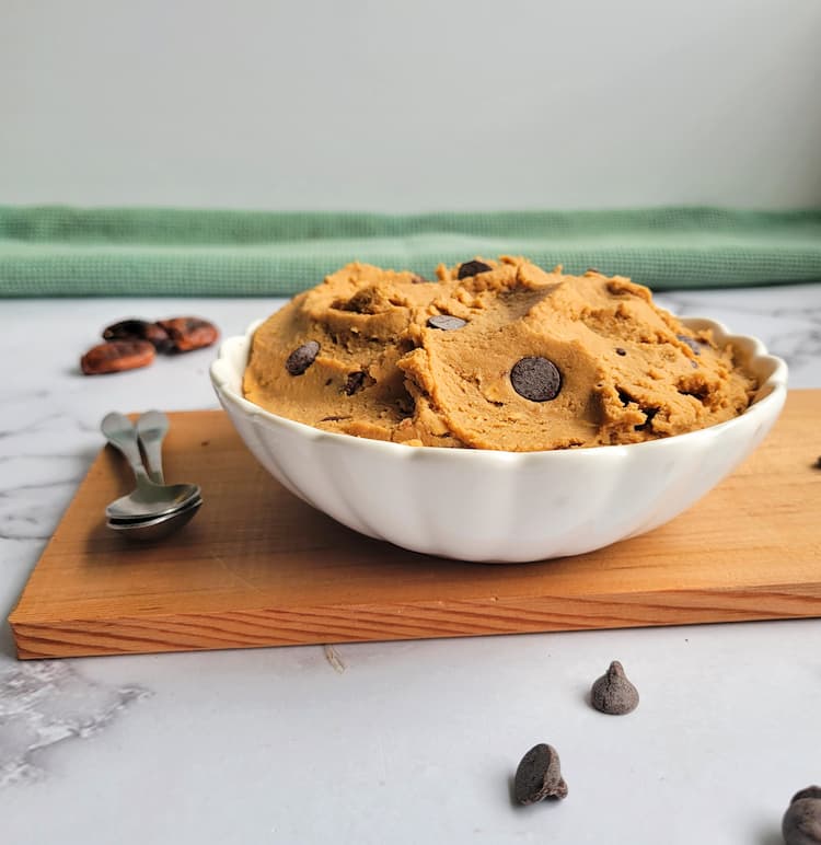 side view of a bowl of chickpea cookie dough with chocolate chips, dates in the background, spoons on the side