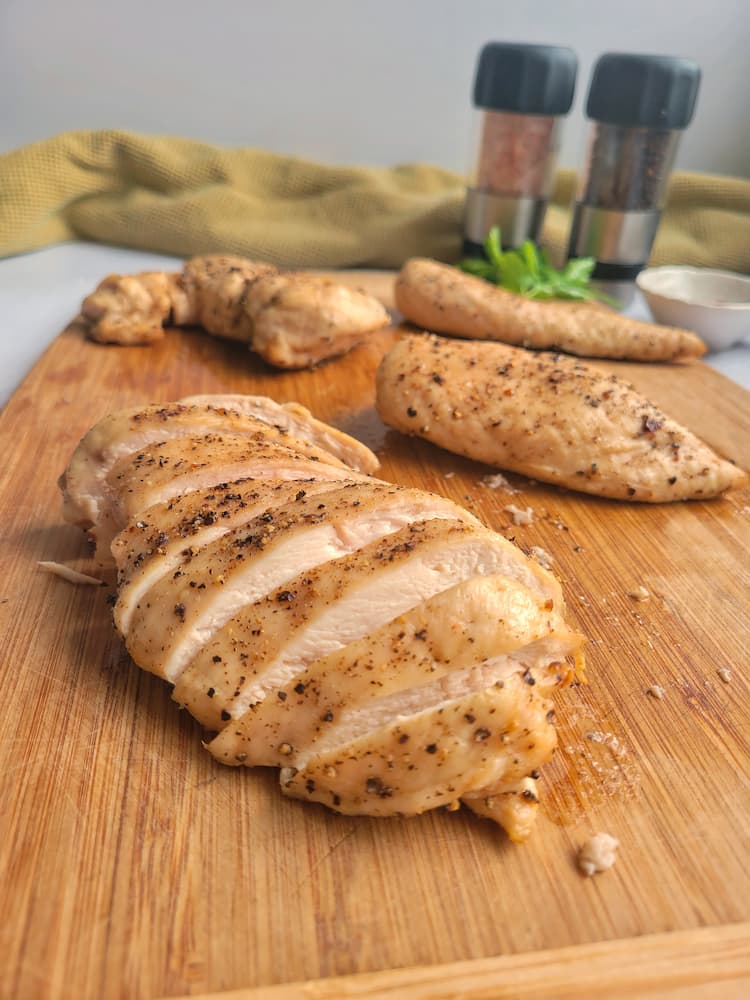 4 chicken breasts on a cutting board, the front one sliced up, salt and pepper shakers in the background
