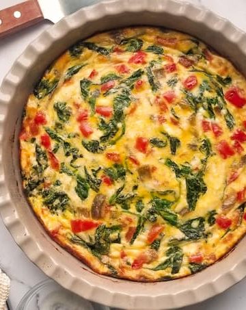 baked quiche with red peppers and spinach in a pie dish, cup of coffee, serving spatula, plates and forks and small ramekin of chili flakes in the background
