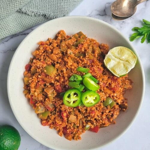 bowl of cauliflower rice mexican, half a lime in the bowl with sliced green onions and jalapenos, whole lime at the bottom, two spoons and cilantro in the background
