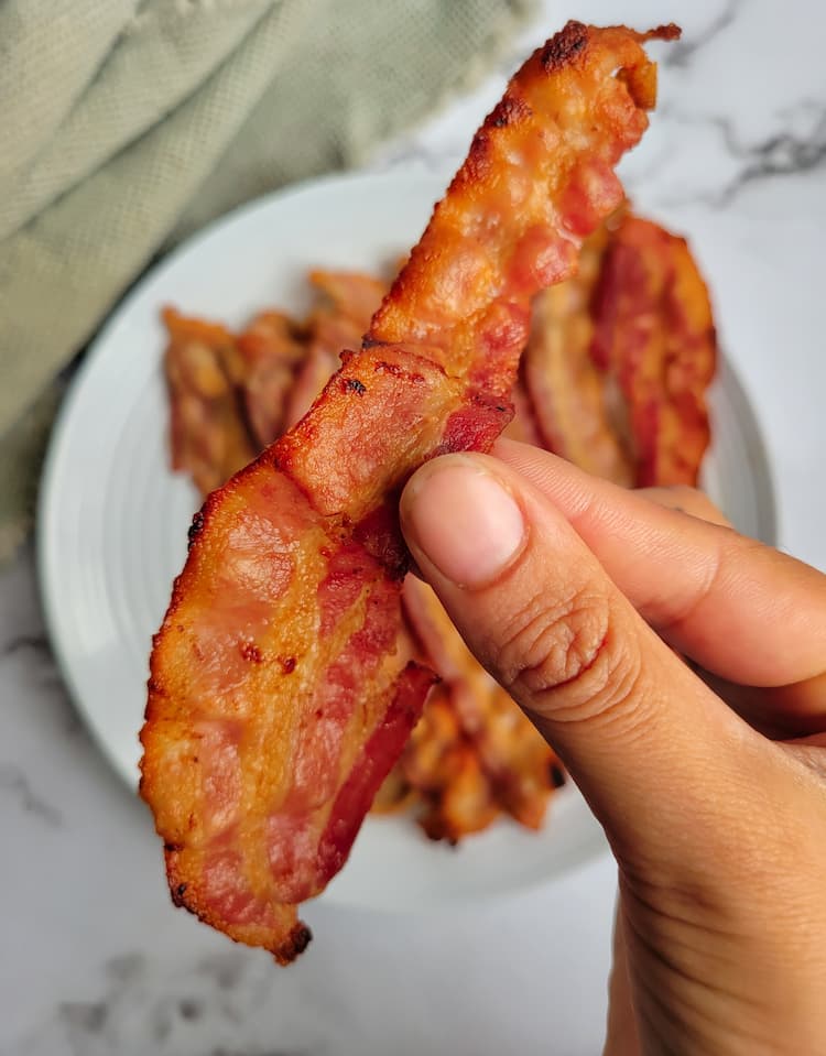 hand holding up a cooked piece of bacon over a plate of the rest