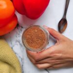 hand holding a jar of homemade fajitas seasoning next to a wooden spoon, red and orange pepper in the background