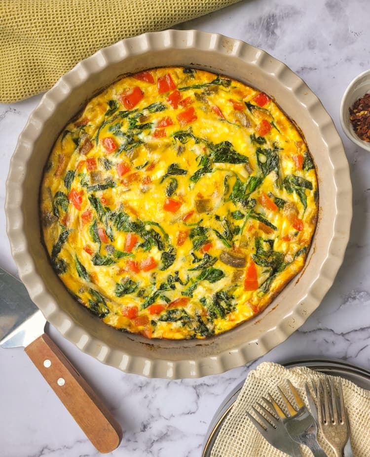 baked quiche with red peppers and spinach in a pie dish, ramekin of chili flakes on the side, serving spatula and plates/forks at the bottom