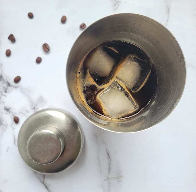 martini shaker with ice and dark drink next to some scattered coffee beans