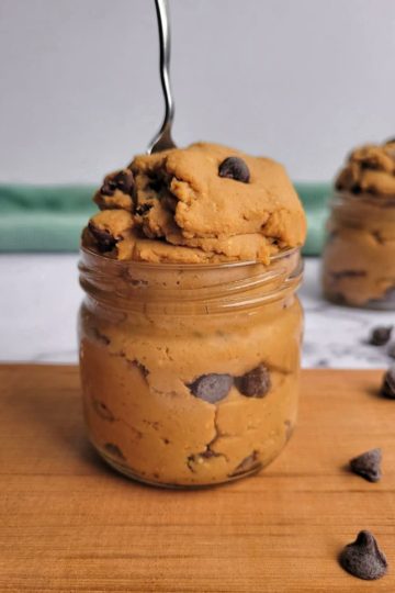 a jar of chickpea cookie dough with chocolate chips, spoon in the jar, chocolate chips scattered on the side, a second jar of the same thing in the backgorund