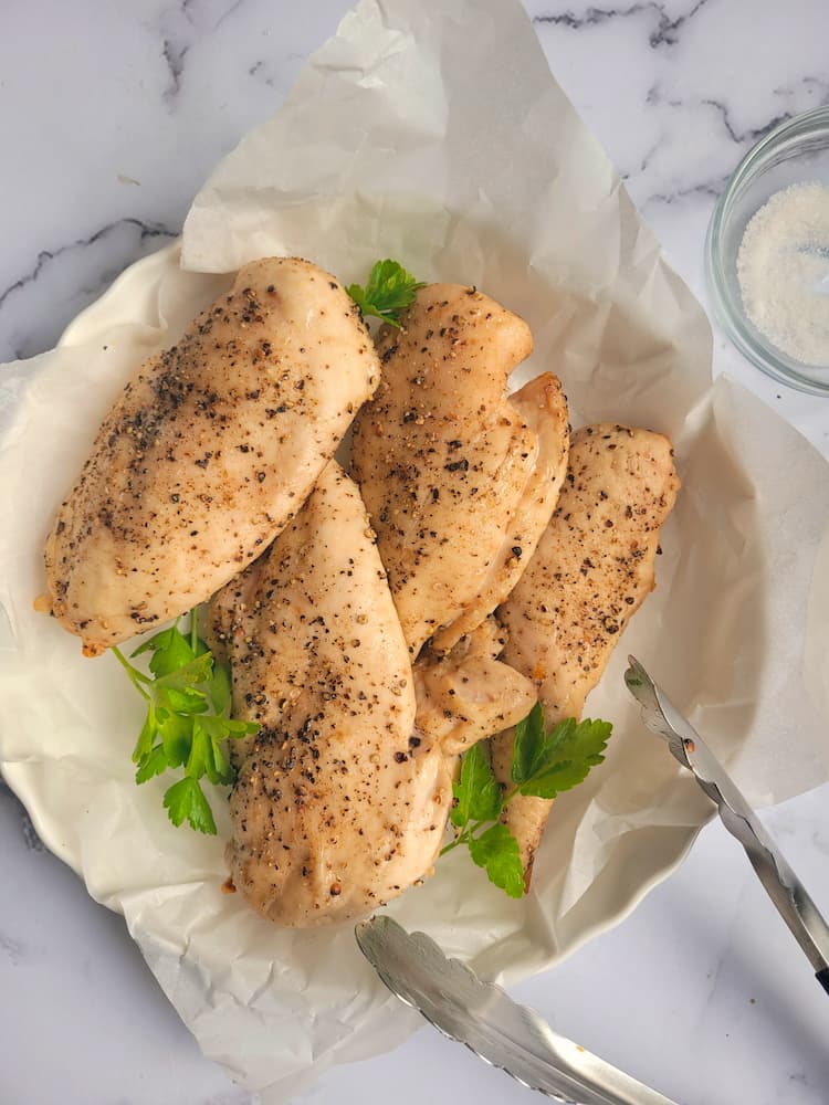parchment lined plate with 4 cooked chicken breasts seasoned with salt and pepper, garnished with parsley leaves, tongs on the side with a small ramekin of salt
