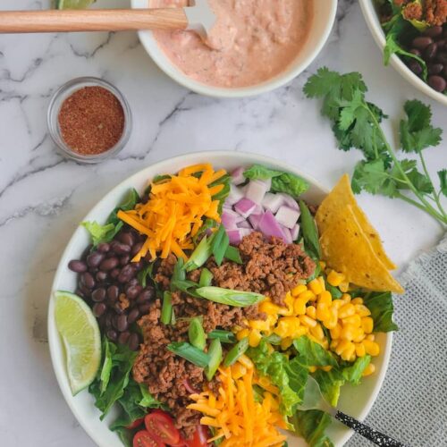 big bowl of taco salad with ground beef, beans, cheese, corn, onions, lettuce, tomatoes, lime wedges, ramekin of taco seasoning in the background with a bowl of sour cream and salsa