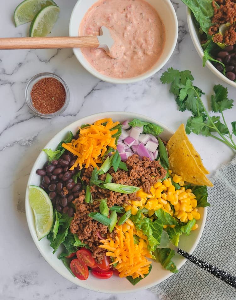 big bowl of taco salad with ground beef, beans, cheese, corn, onions, lettuce, tomatoes, lime wedges, ramekin of taco seasoning in the background with a bowl of sour cream and salsa