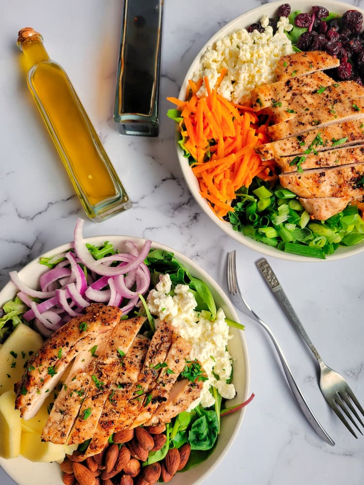 two salads with grilled chicken next to a bottle of oil and vinegar, two forks at the bottom