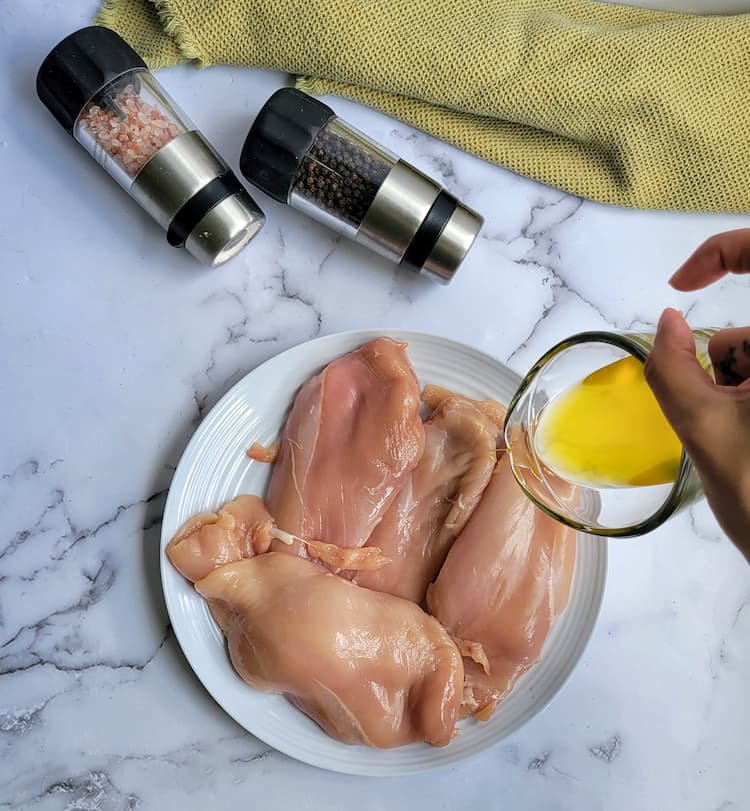 hand pouring a cup of olive oil over a plate of raw chicken breasts, salt and pepper grinders in the background