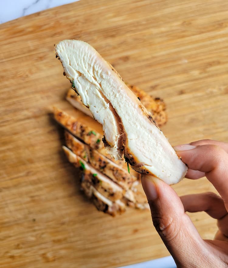 hand holding up a slice of a grilled chicken breast, the rest of the breast in on a cutting board underneath