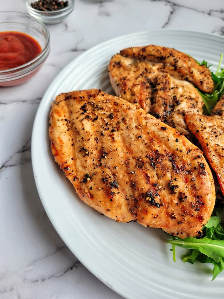 plate with a couple grilled chicken breasts seasoned with salt and pepper, bowl of sauce in the background beside a small ramekin of peppercorns