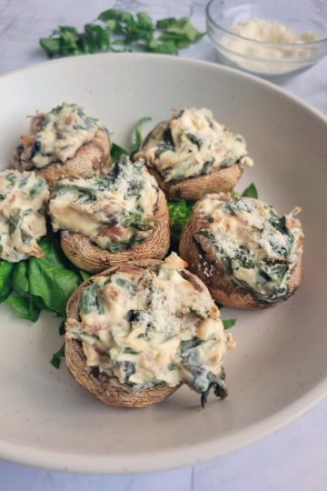 side view of 6 creamy stuffed mushrooms on a plate, ramekin of parmesan cheese in the background next to some fresh herbs