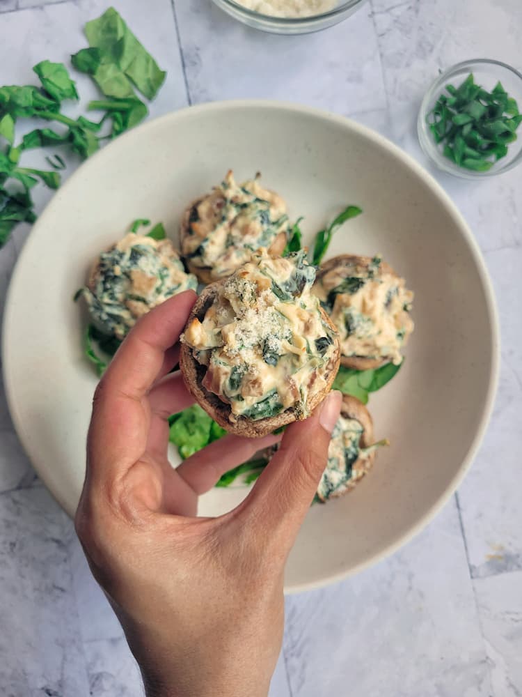 hand holding up a stuffed mushroom over a plate with the rest, ramekin of sliced green onions and fresh herbs in the background