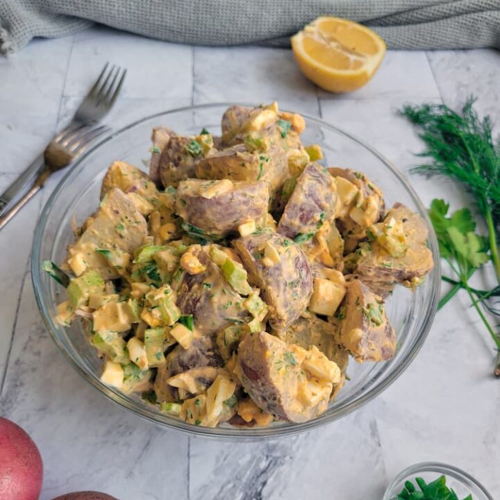 bowl of creamy potato salad with hard boiled egg, celery and herbs,, halved lemon and fresh dill on the side with two forks, bowl of green onions at the bottom next to 2 red potatoes