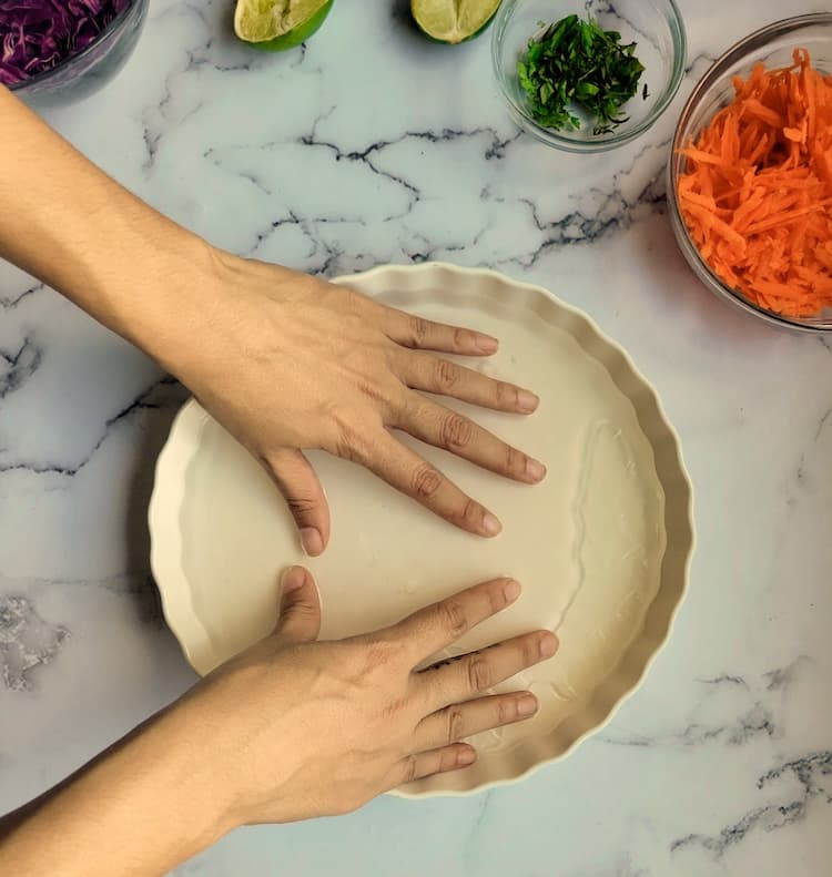 hand pushing down a rice paper into a bowl of water, next to a bowl of shredded carrots, halved limes, chopped parsley and shredded red cabbage