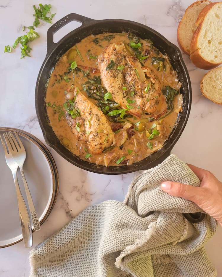 hand holding a tea towel wrapped handle on a cast iron skillet with creamy tuscan chicken, sliced bread in the background, 2 forks and plates on the side