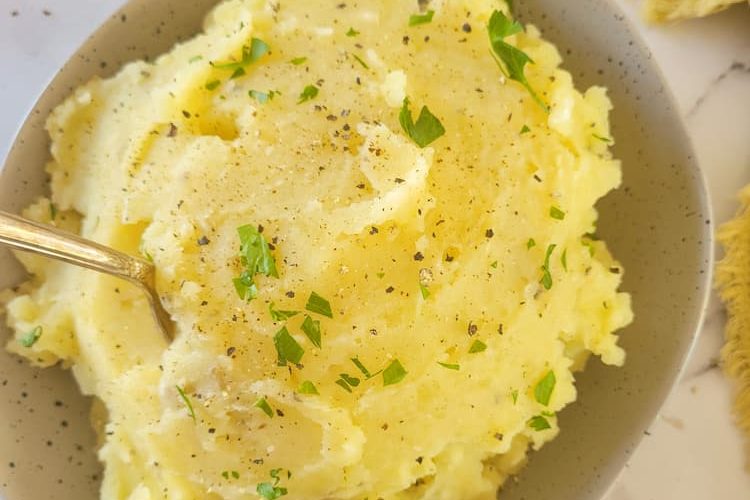 close up of a bowl of mashed potatoes with a spoon, fresh herbs on top and in the background