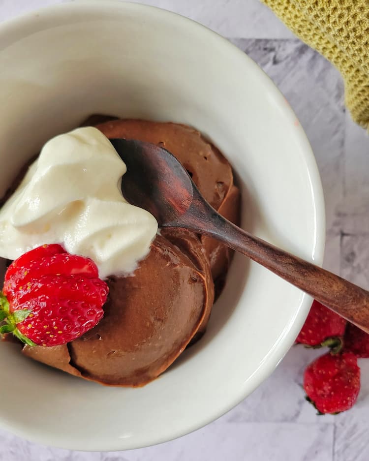 bowl of chocolate mousse made from avocado with whipped cream and a strawberry on top, wooden spoon inside, more strawberries in the background