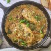 cast iron skillet with 2 chicken breasts in a creamy sauce with fresh herbs, olives and sundried tomatoes, sliced bread and chopped herbs in the background