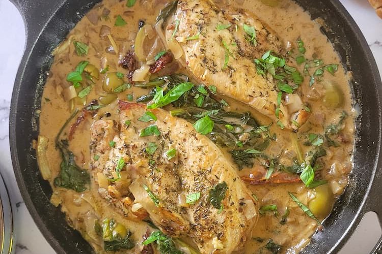 cast iron skillet with 2 chicken breasts in a creamy sauce with fresh herbs, olives and sundried tomatoes, sliced bread and chopped herbs in the background