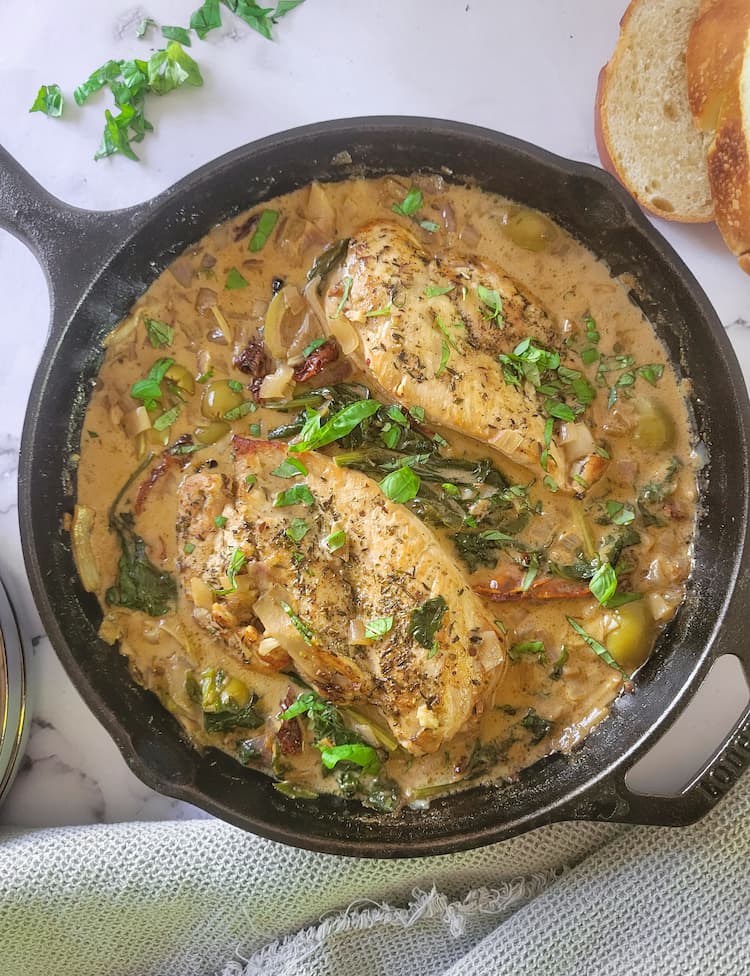 cast iron skillet with 2 chicken breasts in a creamy sauce with fresh herbs, olives and sundried tomatoes, sliced bread and chopped herbs in the background