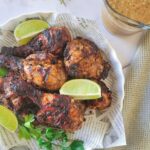 newspaper lined plate with jerk chicken pieces garnished with fresh lime wedges and parsley, jar of jerk sauce in the background
