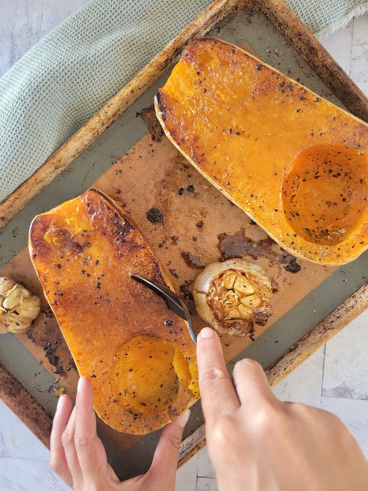 baking sheet with two roasted butternut squash halves and two roasted garlic bulbs, hand holding a spoon scooping the flesh out of one