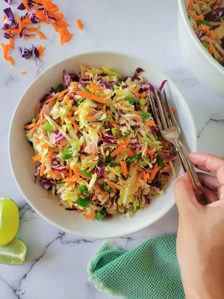 bowl of coleslaw with 2 forks, hand touching the forks, shredded cabbage and carrots and sliced lime in the background with another bowl of coleslaw