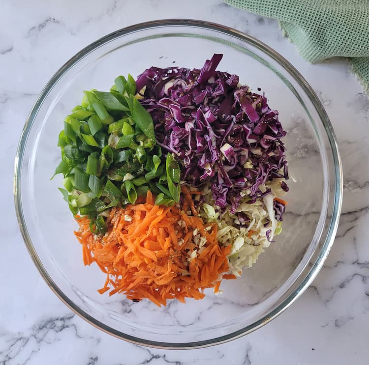 large bowl with chopped green onions, grated carrots, shredded red and green cabbage with dressing drizzled on top