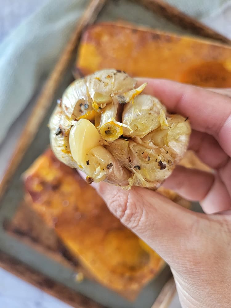 hand holding up a bulb of roasted garlic over a sheet pan with roasted butternut squash
