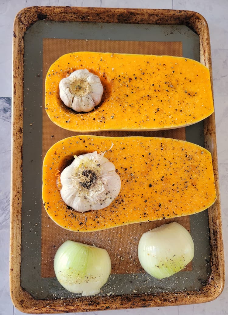 sheet pan with two halves of a butternut squash with whole garlic bulbs in each cavity and a halved onion, seasoned with salt and pepper