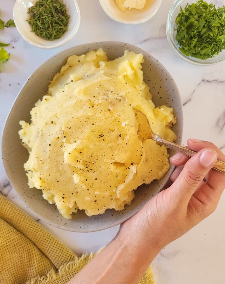 hand holding a spoon in a bowl of garlic mashed potatoes, bowls of fresh herbs and butter in the background