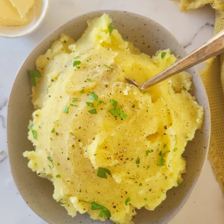 bowl of garlic mashed potatoes with a spoon, garnished with fresh parsley and black pepper, ramekin of butter in the background