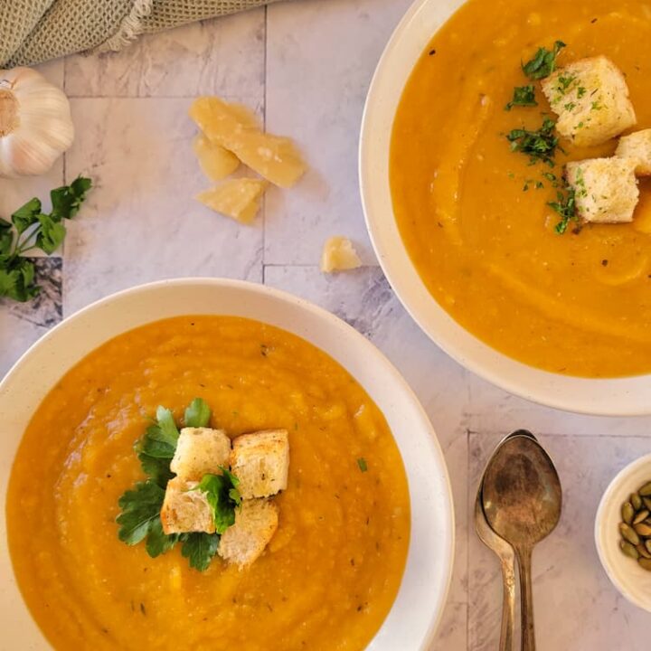 two bowls of butternut squash soup garnished with parsley and croutons, two spoons on the side with parmesan cheese chunks, a garlic bulb and fresh herbs