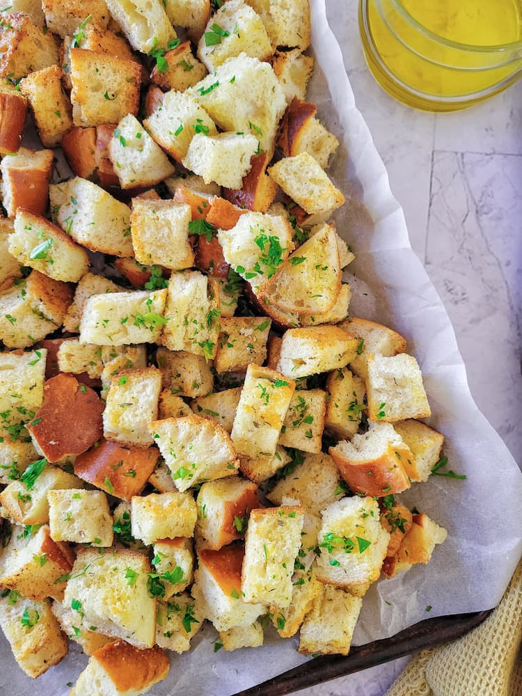 parchment lined baking sheet with a pile of croutons garnished with fresh chopped parsley, jar of olive oil in the background