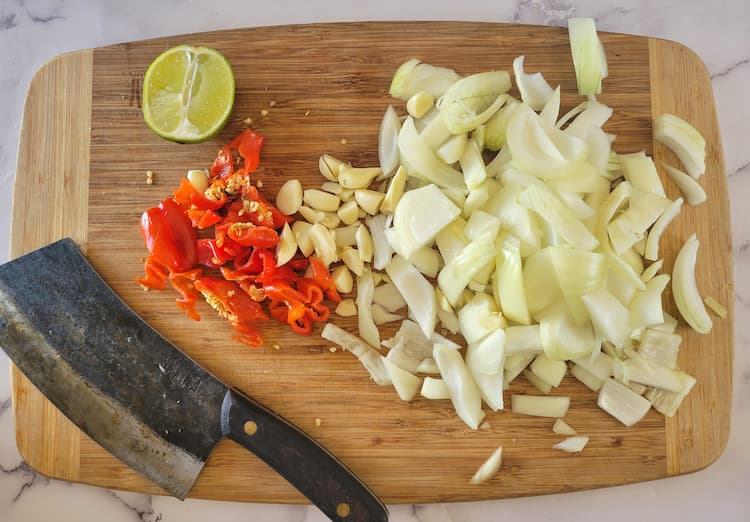 cutting board with chopped onions, garlic, habaneros and a half of a lime with a big knife